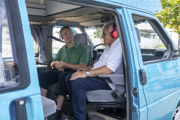 A young man and an elderly man in a wheelchair share a joyful moment inside a blue camper van. The younger man, seated in the driver's seat, is attentively listening to the elderly man who is wearing red headphones
