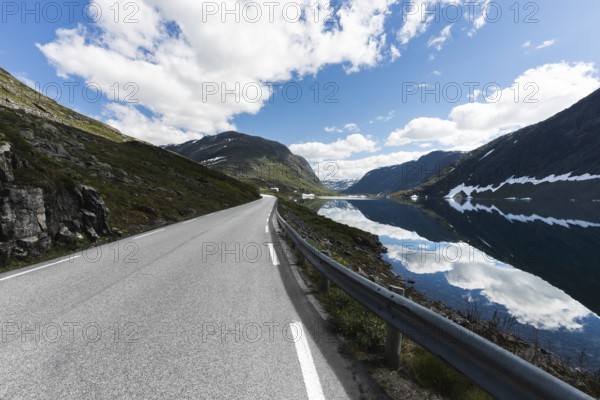 A stunning road in Norway stretches into the distance, flanked by majestic mountains and a mirror-like lake The clear blue sky enhances the outdoor trekking and camping vibe