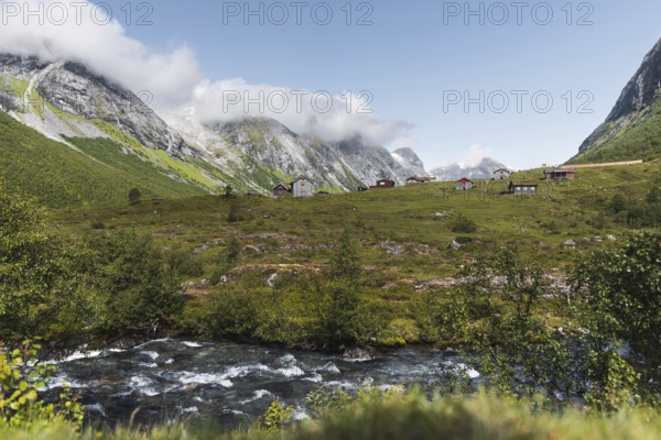 A serene mountain village nestled in a lush green valley in Norway, surrounded by towering peaks and a flowing river Misty clouds add a magical touch to this idyllic landscape
