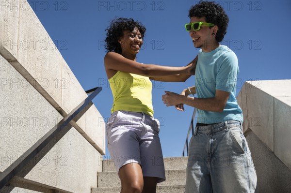A vibrant, multiethnic LGBTQ+ couple shares a joyful moment on outdoor steps. Both dressed in casual summer attire, they express happiness under a clear blue sky