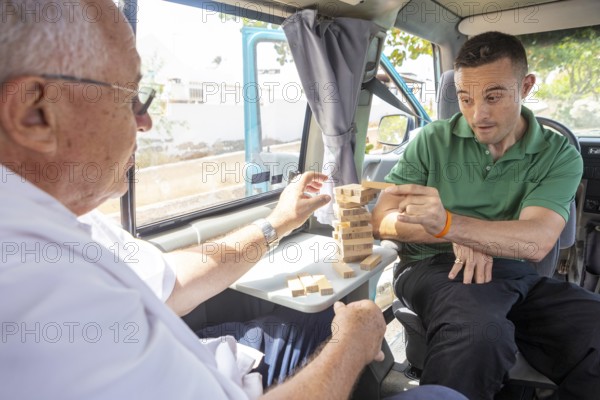 An elderly man and his adult son enjoy playing a game of Jenga inside a van, parked in a sunny suburban area. The elderly man is in a wheelchair, showing accessible van features