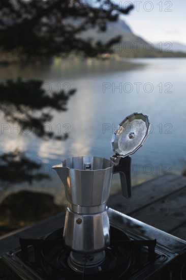A moka pot sits on a camping stove beside a tranquil Norwegian lake, surrounded by lush mountains Perfect setting for a relaxing outdoor adventure and coffee lovers