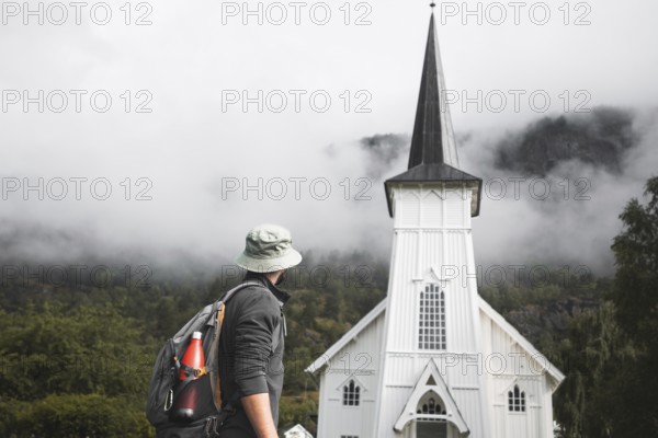 A traveler with a backpack and hat stops in front of a picturesque white Norwegian church, surrounded by mist-covered mountains and lush greenery, capturing the essence of a road trip through Norway
