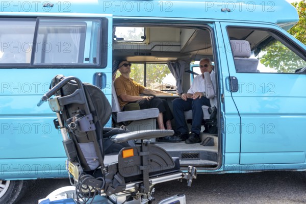 Two men sitting inside a blue van that's adapted for wheelchair access, with the wheelchair positioned outside