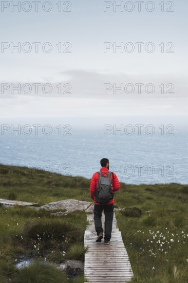 A traveler wearing a red jacket and backpack walks along a wooden path overlooking the Norwegian sea The lush greenery and vast ocean create a perfect scene for an adventurous roadtrip