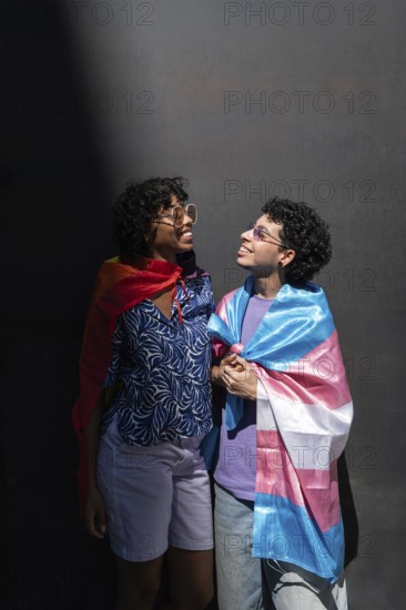 A joyful multiethnic LGBTQ+ couple smiling at each other, wrapped in Pride flags. Captured in natural light against a textured background, exuding love and acceptance