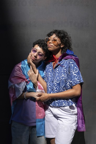 A multiethnic LGBTQ+ couple stands closely together, wrapped in colorful flags. Both wear sunglasses and embrace under a sunlit backdrop, expressing love and pride
