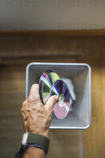 A person hand dropping assorted DVDs into a trash bin. The image highlights the decline of physical media formats in the digital age, capturing a recycling concept