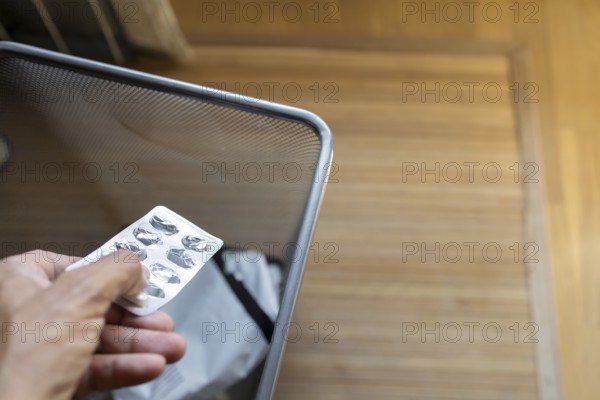 A hand discarding an pill blister pack into a mesh waste bin in a room with a wooden floor. The image depicts themes of waste disposal and medication use
