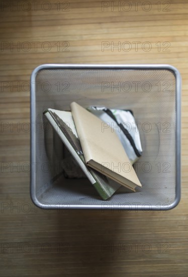 Aerial shot of a metal mesh waste bin containing discarded books, placed on a wooden floor. The image evokes themes of waste, recycling, and environmental consciousness