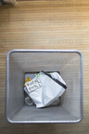A top down view of a trash bin containing used pill packaging and other waste, set on a wooden floor. This image highlights daily household waste disposal