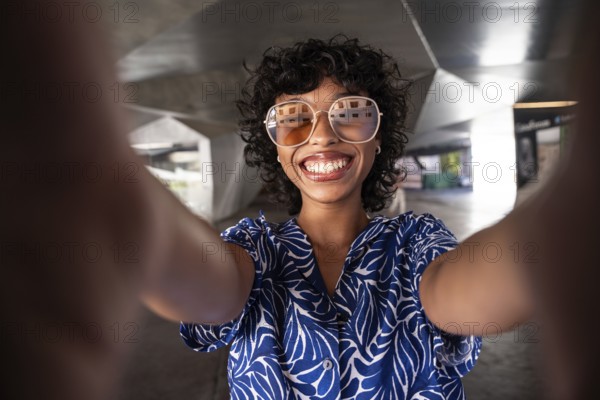Happy woman with curly hair and sunglasses takes a selfie in an industrial setting. She wears a blue patterned shirt and smiles brightly at the camera