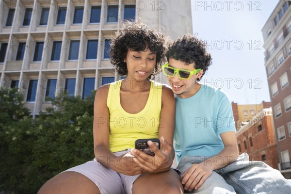 A multiethnic LGBTQ+ couple enjoys a sunny day in the city, looking at a smartphone together. The backdrop features modern architecture and urban vibes, capturing a lively moment