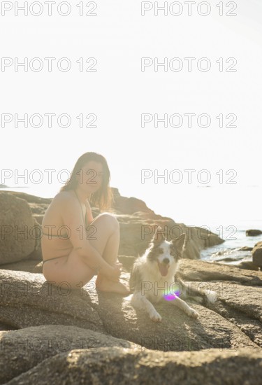 A woman in a bikini sits beside her happy dog on sunlit rocks by the sea The peaceful setting suggests relaxation and a love for nature and pets