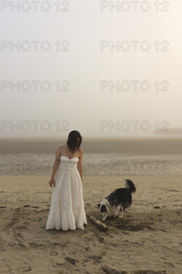 A woman in a white dress stands on a misty beach with a playful dog nearby The serene atmosphere and soft colors evoke calmness and a sense of peace