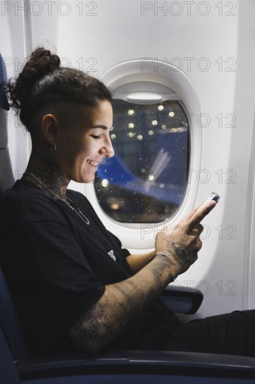 A young woman with intricate tattoos sits on an airplane, smiling while using a smartphone The window reveals a beautiful night, and the ambient light creates a relaxed atmosphere
