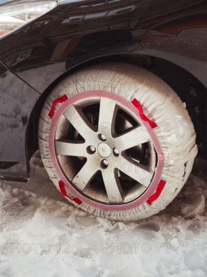 A close-up of a car's wheel equipped with a red snow chain, partially covered in snow, highlighting winter urban driving conditions