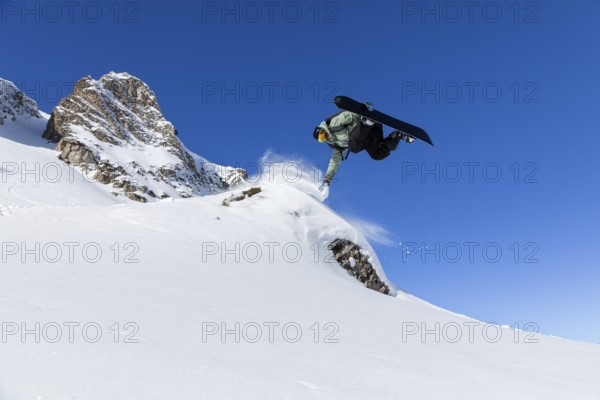 A snowboarder executes an aerial trick on a snowy slope under a vibrant blue sky. The dynamic movement captures the thrill and skill of winter sports in a mountainous landscape