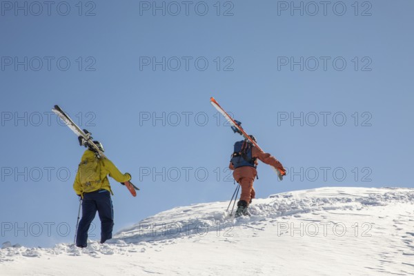 Two skiers in colorful gear hike up a snowy slope, carrying their skis The clear blue sky contrasts with the pristine snow, capturing the thrill of alpine freeride skiing