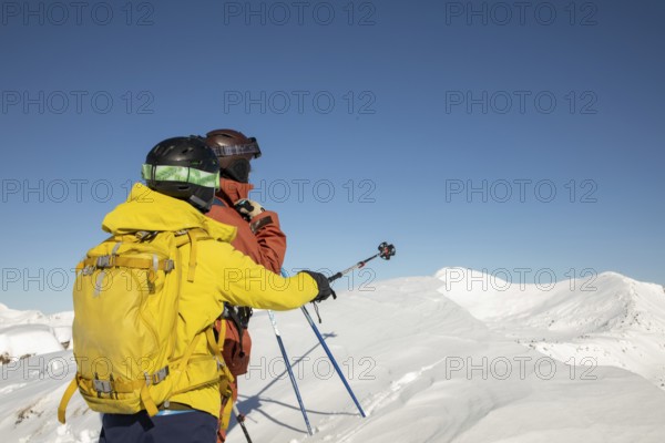 Two freeride skiers stand atop a snow-covered peak, gazing into the vast mountain range under a clear blue sky Equipped with helmets and colorful gear, ready for adventure