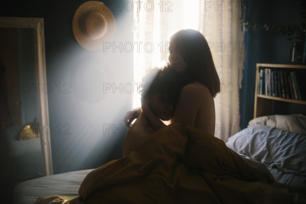 A couple sits on a cozy bed, enveloped in a peaceful embrace. Soft morning sunlight streams through curtains, casting a warm glow in the intimate, tranquil bedroom setting