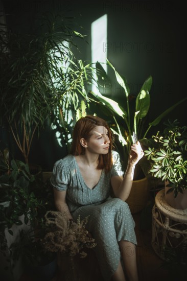 A woman in a green dress sits surrounded by lush indoor plants, holding a small card. Sunlight filters through the leaves, creating a serene atmosphere