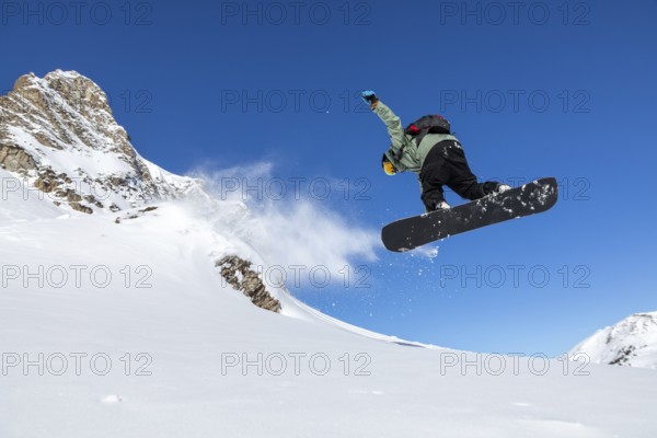 A snowboarder performs a jump against a clear blue sky, with snow spraying behind. The stunning mountain backdrop adds to the thrill of the high speed winter sport action