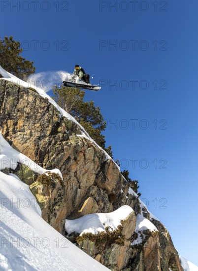 A snowboarder performs an impressive jump off a snow covered cliff, showcasing athleticism and adventure against a clear blue sky backdrop. The thrill of winter sports comes alive