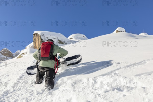 A snowboarder, wearing a red backpack, climbs a snowy mountain with a snowboard. The scene captures adventure, determination, and the thrill of winter sports