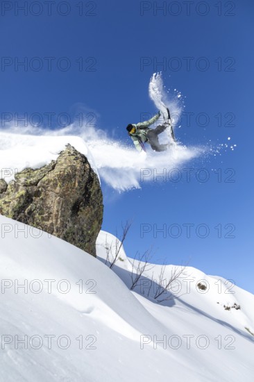 A snowboarder performs an aerial trick off a snowy cliff against a clear blue sky, capturing the thrill and excitement of winter sports in a pristine mountain setting