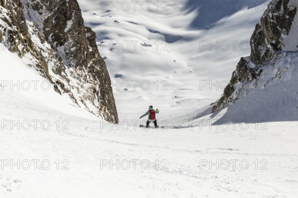 A snowboarder carves down a steep, snow covered slope between rocky peaks. The bright snow contrasts with the rugged mountain, creating a scene of adventure and thrill