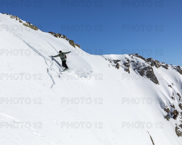 A snowboarder skillfully descends a steep, snowy slope against a clear blue sky. The untouched snow and rocky cliff create a stunning winter sports scene