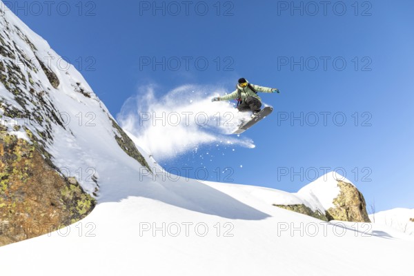 A snowboarder executes an impressive jump, sending a spray of snow flying. The scene is set against a vibrant blue sky and snow covered mountains, capturing the thrill of winter sports