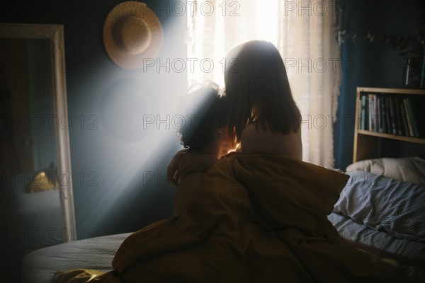 A couple shares an intimate embrace on a bed, bathed in warm morning light streaming through a window. The cozy room setting includes a mirror, straw hat, and bookshelf