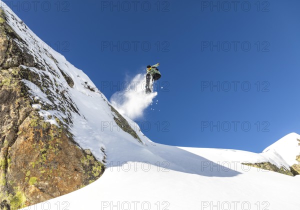 A snowboarder performs an impressive jump off a snow covered mountain, leaving a trail of snow against a vibrant blue sky, capturing the thrill of alpine adventure