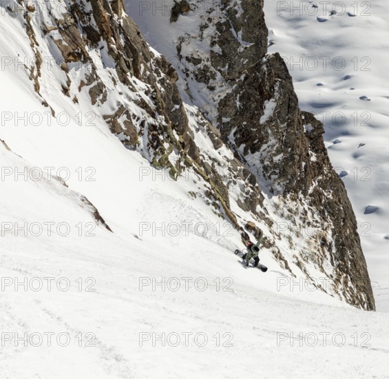 A snowboarder skillfully descends a steep, snowy slope surrounded by rugged cliffs and untouched snow. The scene captures the thrill and challenge of mountain snowboarding