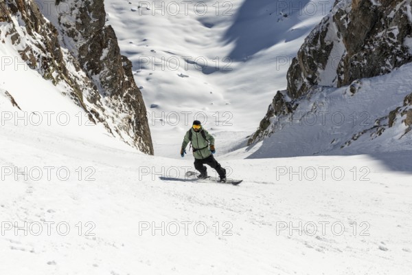 A snowboarder skillfully descends a steep, snowy mountain slope surrounded by rugged cliffs. The scene captures the thrill of winter sports and the beauty of a winter landscape