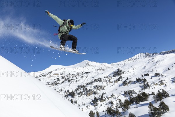 Snowboarder performs a jump on snowy mountain slope, surrounded by a breathtaking winter landscape. The clear blue sky enhances the action and adventure of the scene