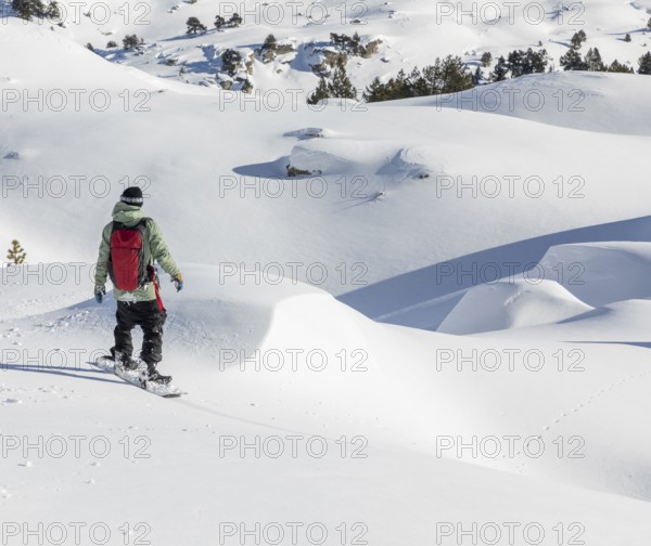 A snowboarder wearing a green jacket and red backpack navigates a pristine snowy landscape, surrounded by untouched snow and distant trees under a clear blue sky