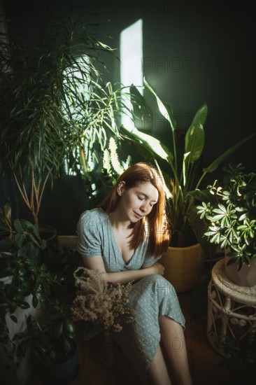 A woman in a light dress calmly sits surrounded by lush indoor plants. Sunlight filters through, casting a serene ambiance. Perfect for concepts of tranquility and nature