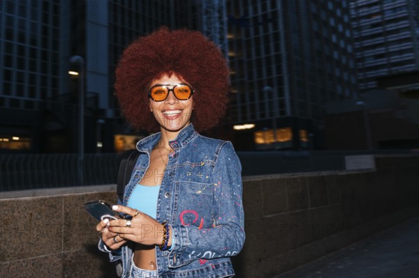 Fashionable African descendent woman with vibrant afro hair smiles while checking her smartphone in a city environment, donning a colorful denim jacket and orange sunglasses