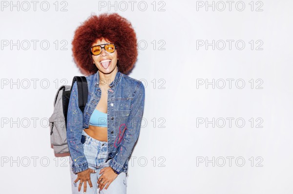 Joyful African descent woman with voluminous afro hair, sticking out her tongue, wearing sunglasses, a denim jacket, and carrying a backpack. She poses against a white background