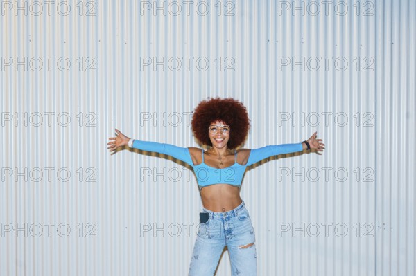 African descendent woman with vibrant afro hair, wearing a bright blue top and jeans, poses joyfully with arms wide open against a ridged metal background