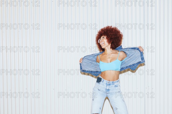 A joyful African descent woman with voluminous afro hair wearing light blue denim jeans, a crop top, and a jacket for a lively pose against a striped background