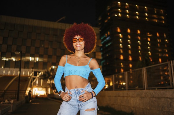 African descendent woman with striking afro hair and sunglasses poses confidently outdoors at night with urban buildings illuminated in the background