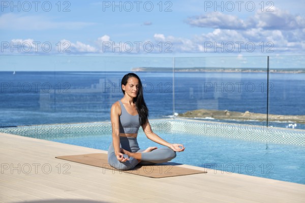 A woman practices yoga in lotus pose beside an infinity pool overlooking the ocean on a terrace. The tranquil scene, with clear skies and calm waters, embodies relaxation and balance