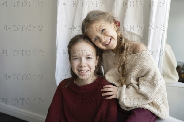 Two sisters wearing cozy sweaters smile warmly at the camera The younger sibling leans affectionately on the other, showing tender family bonding in a comfortable home setting