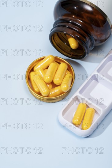 Top view of a dark amber bottle spilling yellow capsules into a gold lid, alongside a white pill organizer on a blue background