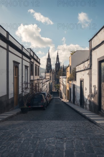 A calm cobblestone street lined with traditional houses leads to the iconic San Juan Bautista Church in Arucas, Gran Canaria. The scene captures a clear sunny day, showcasing the church's intricate Gothic architecture against a bright blue sky
