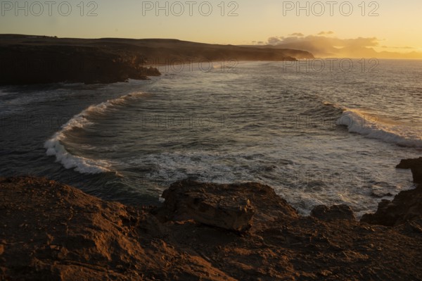 Dramatic view of powerful waves hitting rugged rocks with a sunset backdrop at La Pared, Fuerteventura. The warm golden light enhances the scenic beauty of the coastal landscape